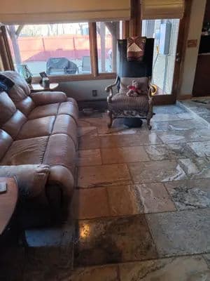 Living room with a leather couch, stone flooring, and a vintage armchair near large windows.