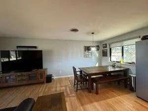 Cozy living room and dining area with wooden table, large TV, and bright windows.