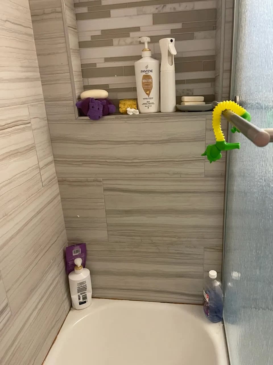 Shower stall with gray tiled walls, shelves containing toiletries, and colorful bath toys.