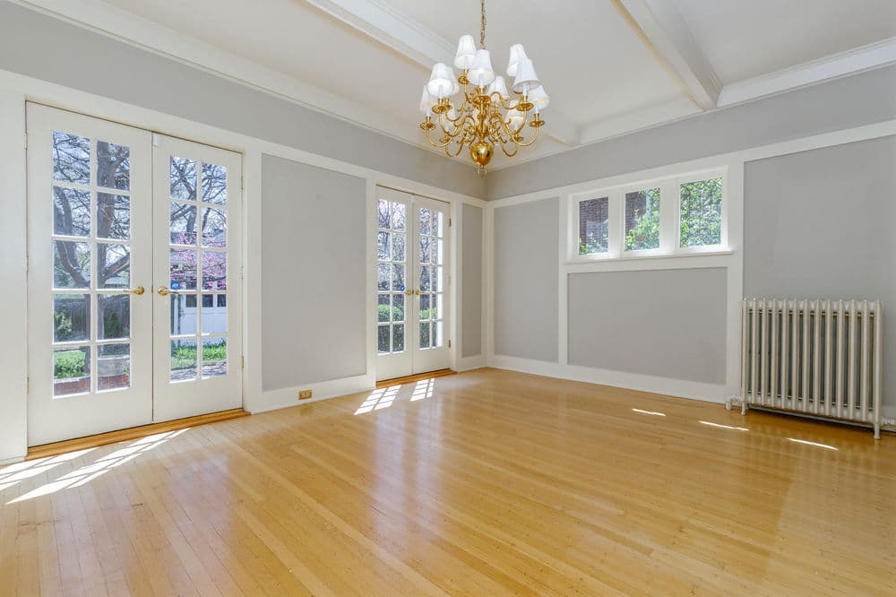 Bright, empty living room with wooden floors, large windows, and elegant chandelier.